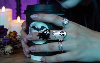 Gothic model hands wearing multiple ghost rings while holding a Halloween themed coffee cup
